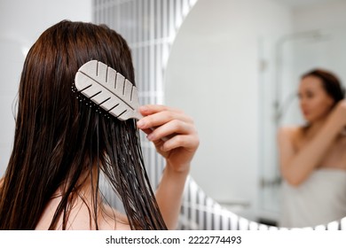 Attractive woman in white towel with comb brushing her wet hair after showering at home in front of bathroom mirror. Cares about healthy and clean hair. Beauty concept