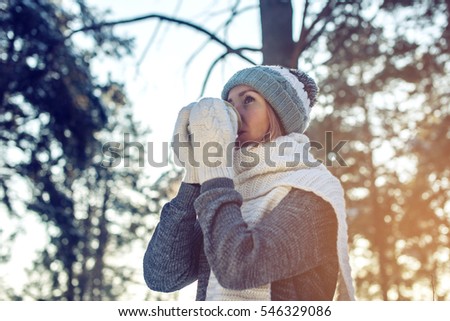 attractive woman in winter, a knitted sweater, mittens and scarf, walking through the Park among the trees and snow in the woods drinking hot tea on a background of sunlight