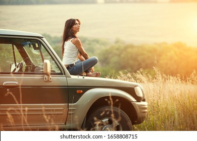 Attractive yong woman is sitting on the car's hood and looking at sunset. Rural evening background.