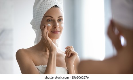 Attractive Young Adult Woman Apply Facial Cream Look In Mirror, Beautiful Healthy Lady Wrapped In Towels Put Moisturizing Lifting Nourishing Day Creme On Soft Hydrated Moisturized Skin In Bathroom