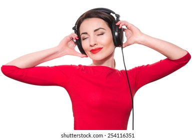 Attractive young adult woman in red clothes listening to the music touching big headphones with closed eyes isolated on white background