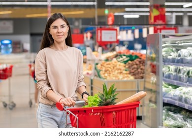 Attractive Young Adult Woman Wearing Casual Clothes Standing With Shopping Cart In Store Aisle