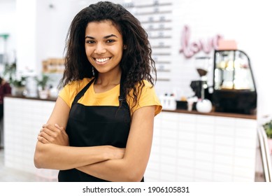 Attractive young African American female barista with curly hair in black apron stands with arms crossed in cafe and friendly smiling. Portrait of pretty waitress wearing uniform - Powered by Shutterstock