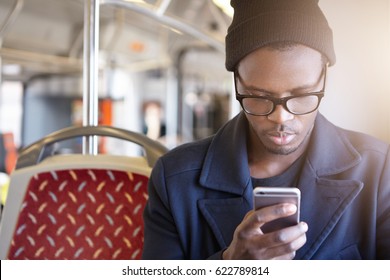 Attractive Young African American Tourist Wearing Black Hat, Stylish Glasses And Coat Using Smart Phone In Bus To Book Airplane Tickets Or Do Online Check In. People, Lifestyle And Modern Technology