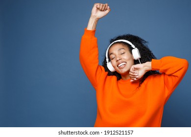 Attractive young african american woman in casual bright orange sweatshirt listening music with headphones dancing rising hands keeping eyes closed isolated on blue color background studio portrait