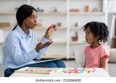 Attractive young african american woman speech therapist working with little black girl, sitting at table with colorful letters, learning pronunciation, clinic interior. Speech disorders in children