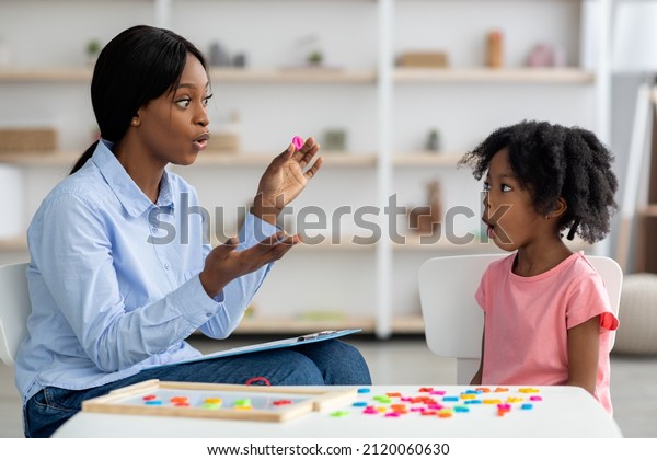 Attractive young african american woman speech\
therapist working with little black girl, sitting at table with\
colorful letters, learning pronunciation, clinic interior. Speech\
disorders in\
children