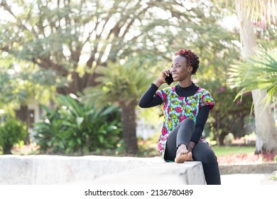 Attractive Young African Girl Sitting In A Park, Radiant With Happiness Talking On Her Cell Phone With Someone She Seems To Care About