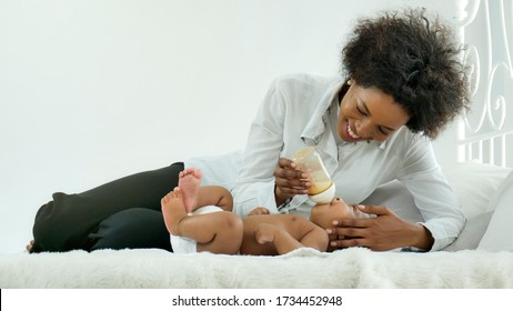 Attractive Young African Woman Feed To Her Baby. Mother Holding And Feeding Baby From Milk Bottle. Loving Woman Giving To Drink Milk On White Bed In The Bedroom.