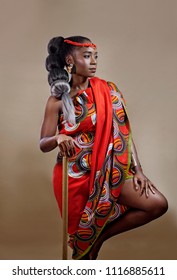 Attractive young African woman with long hair wearing traditional clothing holding a staff while standing on one leg against a brown background