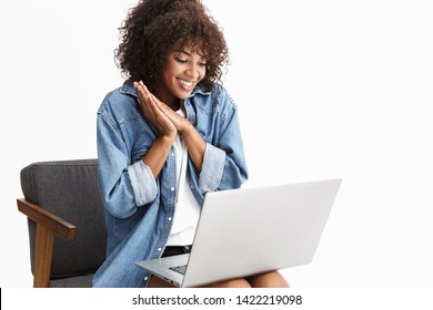 Attractive Young African Woman Wearing Denim Sitting In Chair Isolated Over White Background, Working On Laptop Computer, Chatting