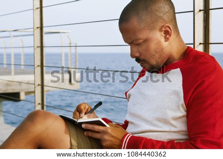 Attractive young African-American hipster man relaxing on the beach and making travel notes in paper notebook