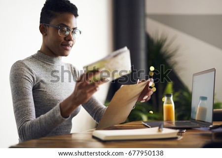 Attractive young afro american businesswoman receiving from post envelope with documentation for project earning money online, professional designer in eyewear packing working papers for mailing 