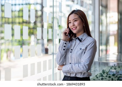 Attraktive junge asiatische Geschäftsfrau, die auf dem Mobiltelefon im Büro steht.