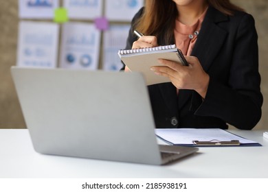 Attractive young Asian business woman in office working on laptop writing work memo on notepad.