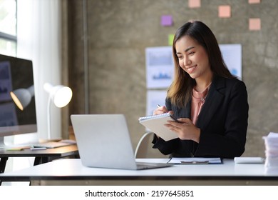 Attractive Young Asian Business Woman In Office Working On Laptop Writing Work Memo On Notepad.