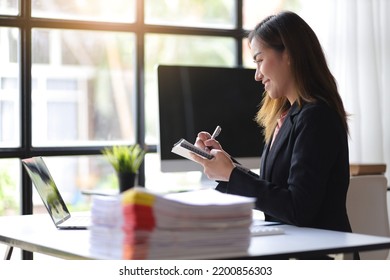 Attractive Young Asian Business Woman In Office Working On Laptop Writing Work Memo On Notepad.