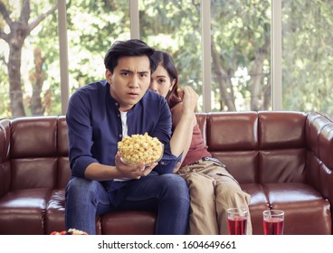 Attractive Young Asian Couple Sitting On Couch In Living Room , Watching Scary Movie,a Man Holding Popcorn In His Hand While His Girlfriend Hiding Behind Him.  