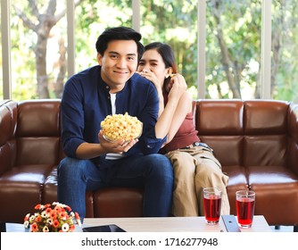 Attractive Young Asian Couple Sitting On Couch , Watching Comedy Movie,a Man Holding Popcorn In His Hand While His Girlfriend Hiding Behind Him And Laughing , Stay Safe  From Covid-19 At Home Concept.