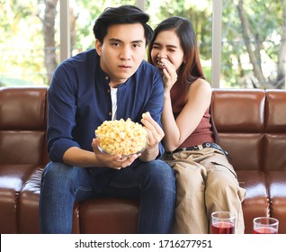 Attractive Young Asian Couple Sitting On Couch , Watching Comedy Movie,a Man Holding Popcorn In His Hand While His Girlfriend Hiding Behind Him And Laughing , Stay Safe  From Covid-19 At Home Concept.