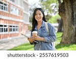 An attractive young Asian female college student in a denim jacket, carrying a backpack and laptop, stands outdoors at her university, smiling at the camera. back to school, education, uni life