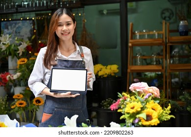 Attractive Young Asian Female Florist Holding And Showing A Digital Tablet Touchpad White Screen Mockup, Standing In Front Of Her Flower Shop.