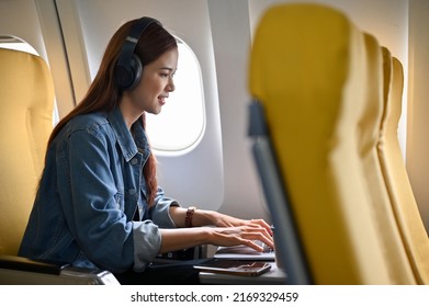 Attractive Young Asian Female Freelancer With Headphones Remote Working On The Plane Sits At The Window Seat Using A Portable Laptop Computer. Vacation Or Journey Concept