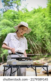 Attractive Young Asian Female Roasting A Pork Chop Beef Steak On A Flying Pan, Cooking With A Portable Picnic Stove. Outdoor Bbq Party Concept