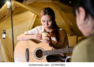 Attractive Young Asian Female Traveler Sipping A Hot Coffee Or Cocoa While Having A Night Camping Party With Her Friends. Teens Camping Concept.