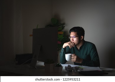 Attractive young asian man drinking coffee sitting on desk table looking at laptop computer in dark late night working feeling serious thinking and determinated at home office in work hard concept.