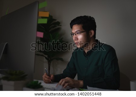 Attractive young asian man sitting on desk table looking at laptop computer in dark late night working feeling serious thinking and determinated at home office in work hard or work load job concept.