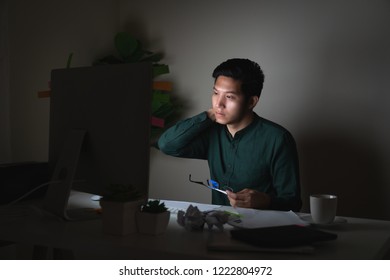 Attractive Young Asian Man Working Late Dark Night Looking At Laptop Computer In Dark Home Office Desk Feeling Tired On Work Load Or Work Hard Concept. Overworked Male Feeling Unhappy And Sleepless.