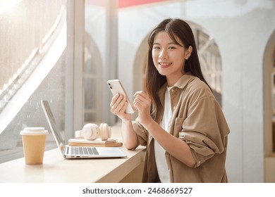 An attractive young Asian woman in casual clothes is holding her smartphone and smiling at the camera while sitting at a table in a coffee shop with her laptop. - Powered by Shutterstock