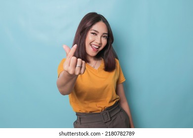 Attractive young Asian woman feels happy and romantic shapes heart gesture expresses tender feelings wears casual yellow t-shirt against blue background. People affection and care concept - Powered by Shutterstock