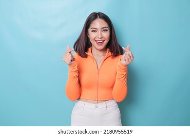 Attractive young Asian woman feels happy and romantic shapes heart gesture expresses tender feelings wears casual orange top against blue background. People affection and care concept