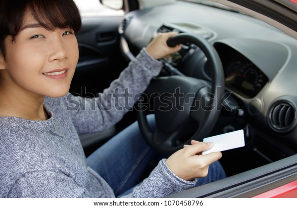 Attractive young asian woman proudly showing\
her drivers license. Smiling woman holding her driver license after\
successful driver\'s exam in her red\
car.
