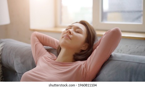 Attractive young asian woman resting on cozy sofa taking deep breath of fresh air holding hands behind head, happy calm mixed race lady relaxing and dreaming on comfortable couch feel stress free