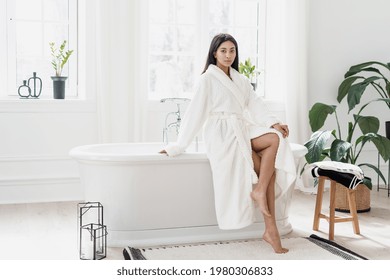 Attractive Young Asian Woman Sitting In Light Spacious Bathroom With Green Plants Wearing White Bathrobe And Showing Legs After Pampering Morning Routine. Selfcare Concept