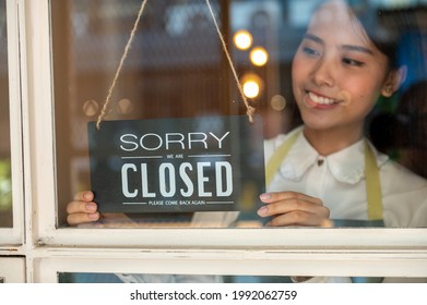 Attractive Young Asian Woman Small Business Owner Hanging Closed Sign On The Glass Window. Focus On Sign. Young Waitress Standing Behind The Door And Turning Closed Sign At The End Of Working Day.