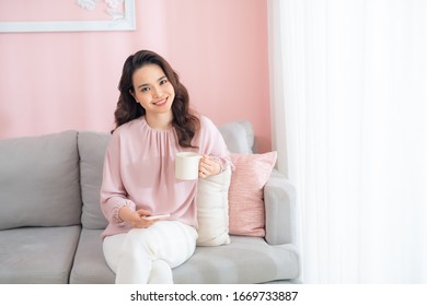 Attractive Young Asian Woman Using Phone And Drinking Coffee When Sitting On Sofa At Home.