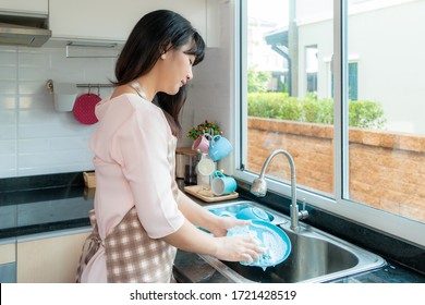 Attractive Young Asian Woman Is Washing Dishes At Kitchen Sink While Doing Cleaning At Home During Staying At Home Using Free Time About Their Daily Housekeeping Routine.
