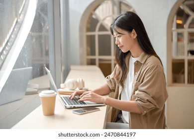 An attractive young Asian woman is working remotely from a coffee shop in the city, working on her laptop computer, typing on the laptop keyboard, responding to emails, working online. - Powered by Shutterstock