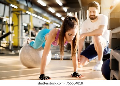 Atractiva joven atlética empujando con el balón de fitness en el moderno gimnasio al lado del apuesto entrenador personal satisfecho.
