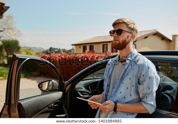 Attractive young bearded male taxi driver in\
sunglasses leaning back on his car with open front door holding\
digital tablet, waiting for client. Unshaven man using portable\
electronic device\
outdoors