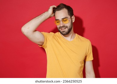 Attractive young bearded man guy in casual yellow t-shirt glasses posing isolated on red wall background studio portrait. People lifestyle concept. Mock up copy space. Looking down, put hand on head - Powered by Shutterstock