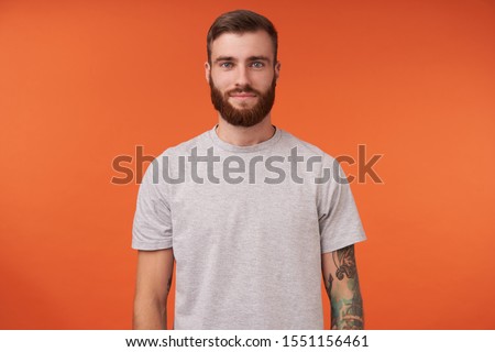 Attractive young bearded tattooed brunette man with trendy haircut standing over orange background, keeping hands along body and looking at camera with charming smile