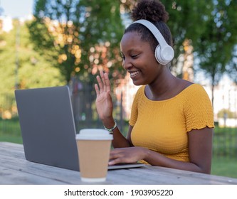 attraktive junge schwarze Frau macht einen Videoanruf in einem Park, verwendet Kopfhörer, grüßt Freunde, entfernte Verwandte mit ihren Händen
