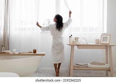 Attractive Young Black Woman Opening Curtains While Making Morning Beauty Routine In Bathroom, Smiling African American Female Wearing White Bathrobe Preparing For Spa Treatments At Home, Back View - Powered by Shutterstock