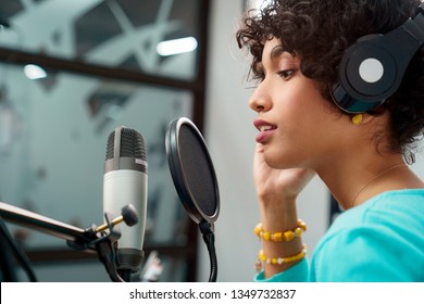 Attractive Young Black Woman Singing Into Microphone in Recording Studio Session. African American Female and podcast host with curly hair in Radio Show Talking to Microphone while wearing headphones - Powered by Shutterstock