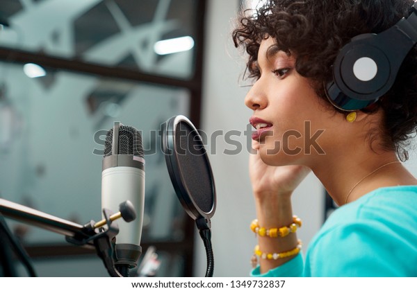 Attractive Young Black Woman Singing Into\
Microphone in Recording Studio Session. African American Female and\
podcast host with curly hair in Radio Show Talking to Microphone\
while wearing\
headphones